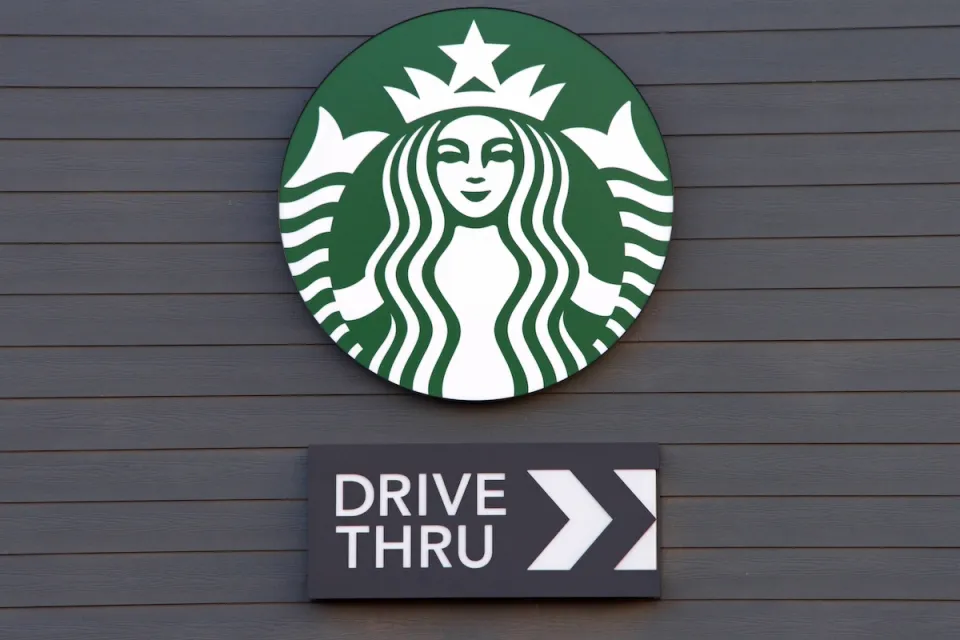 Close-up of Starbucks coffee logo with Drive Thru sign with arrow under it mounted on dark grey wall.